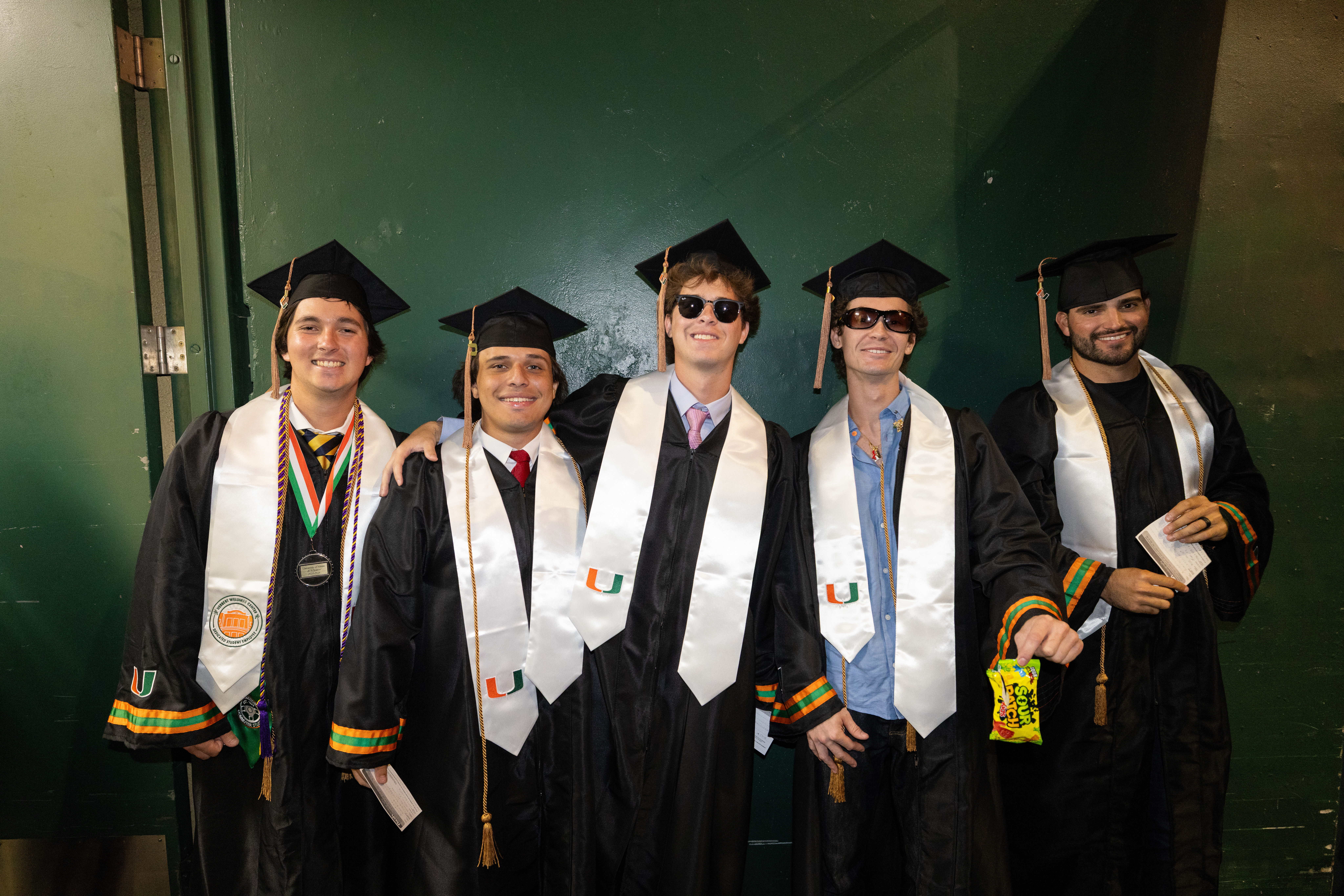 Five smiling graduates in black gowns and caps pose for a photo. The ...