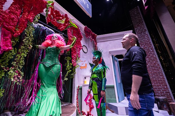 An actor in an elaborate pink puffy wig wearing a bright green costume stands on stage as a man gives her directions. Beside him stands another actor in a bright green costume.