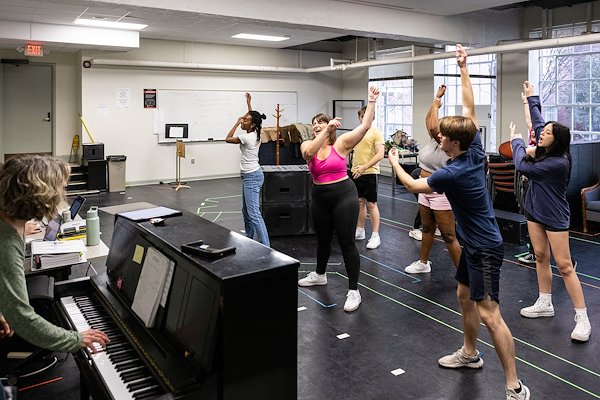 In a room with windows, a blonde woman plays the piano while eight people swing their arms in a dance as they rehearse for a play.