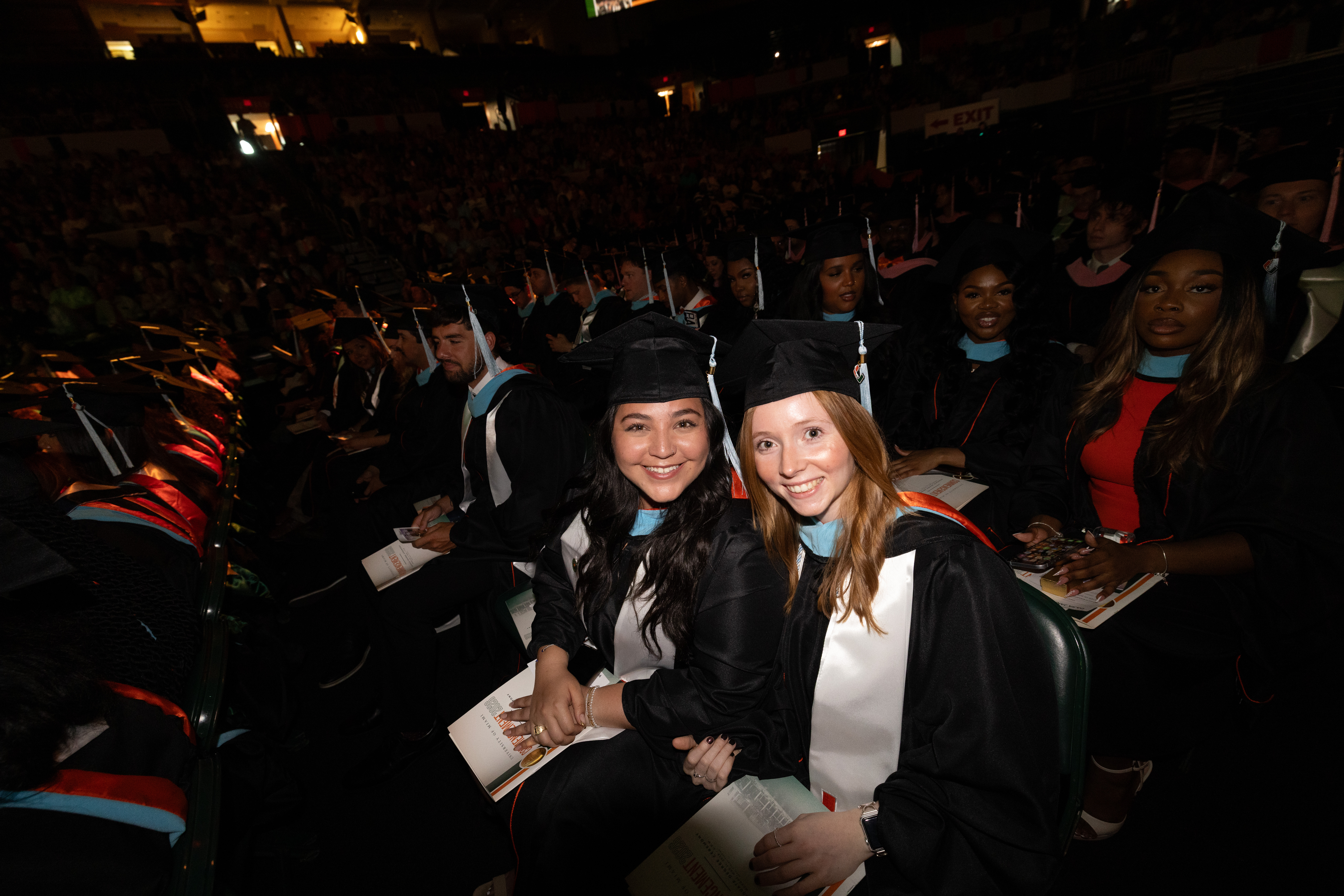 Two smiling graduates in caps and gowns sit in an auditorium during the ...