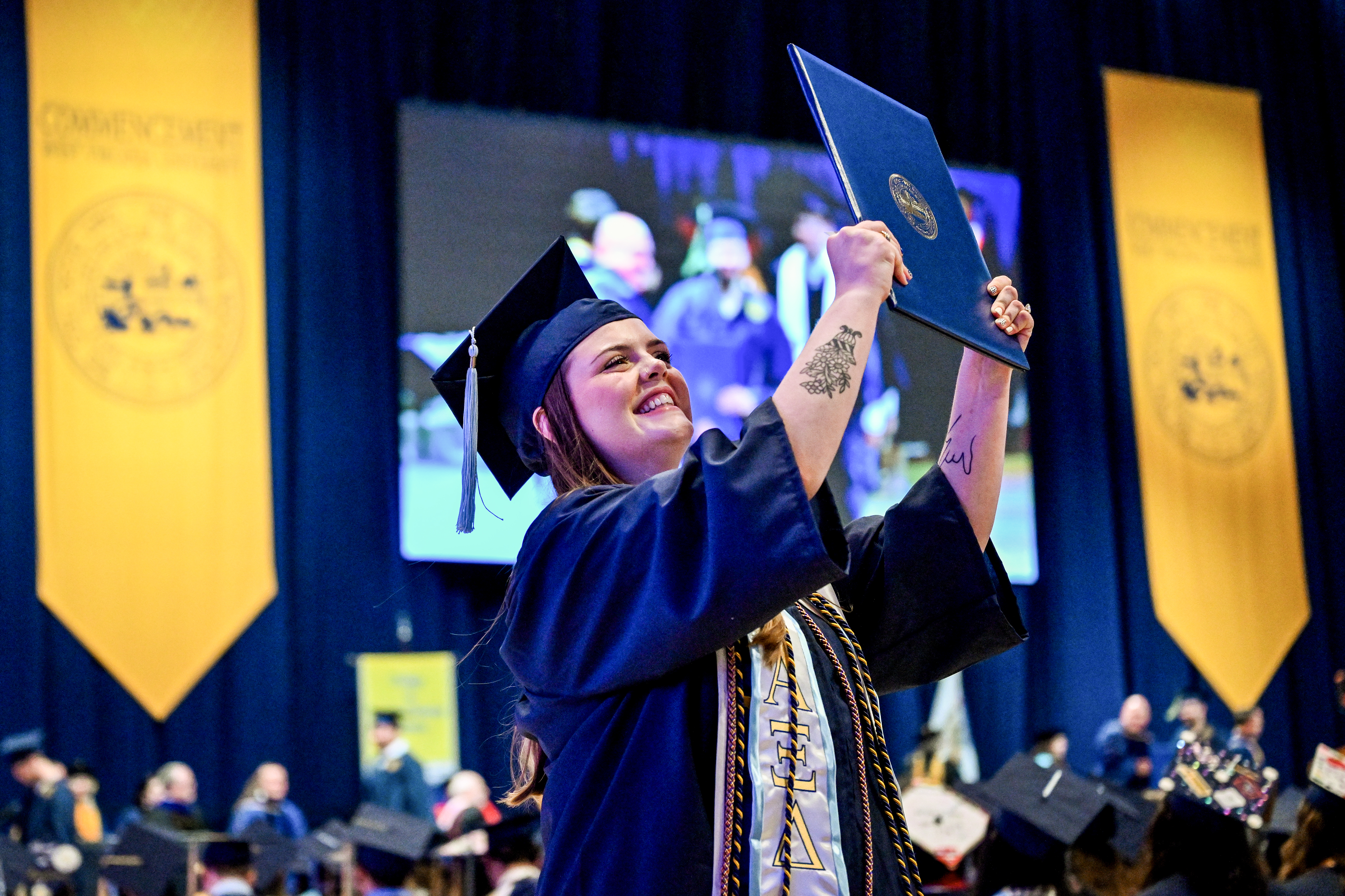 Graduate in cap and gown smiles, holding diploma overhead on stage ...