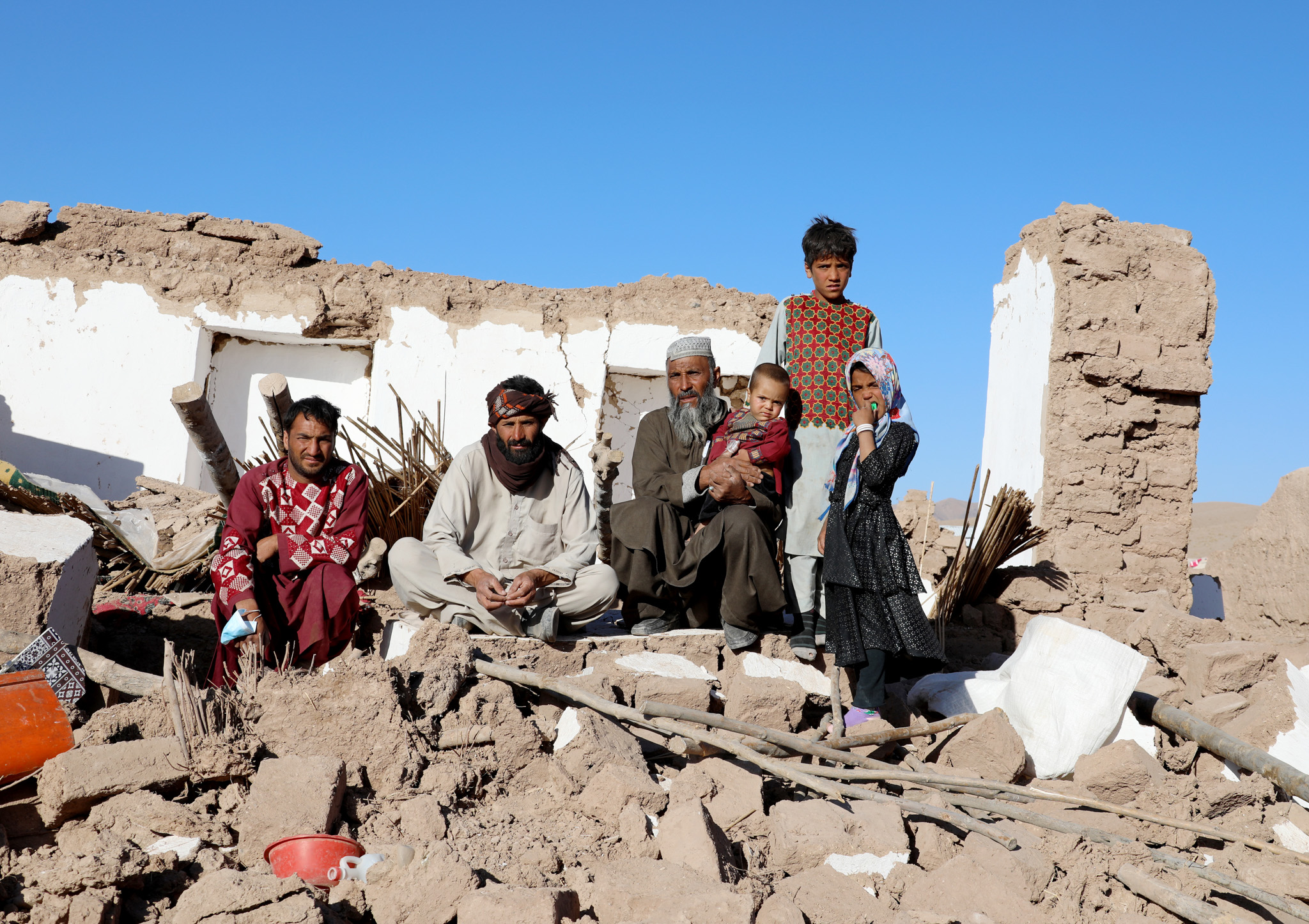 A group of adults and children sit on rubble in front of a partially ...