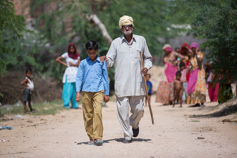 Sankarlal being guided along a dirt track by his granddaughter Priyanka. He carries a staff in his right hand.