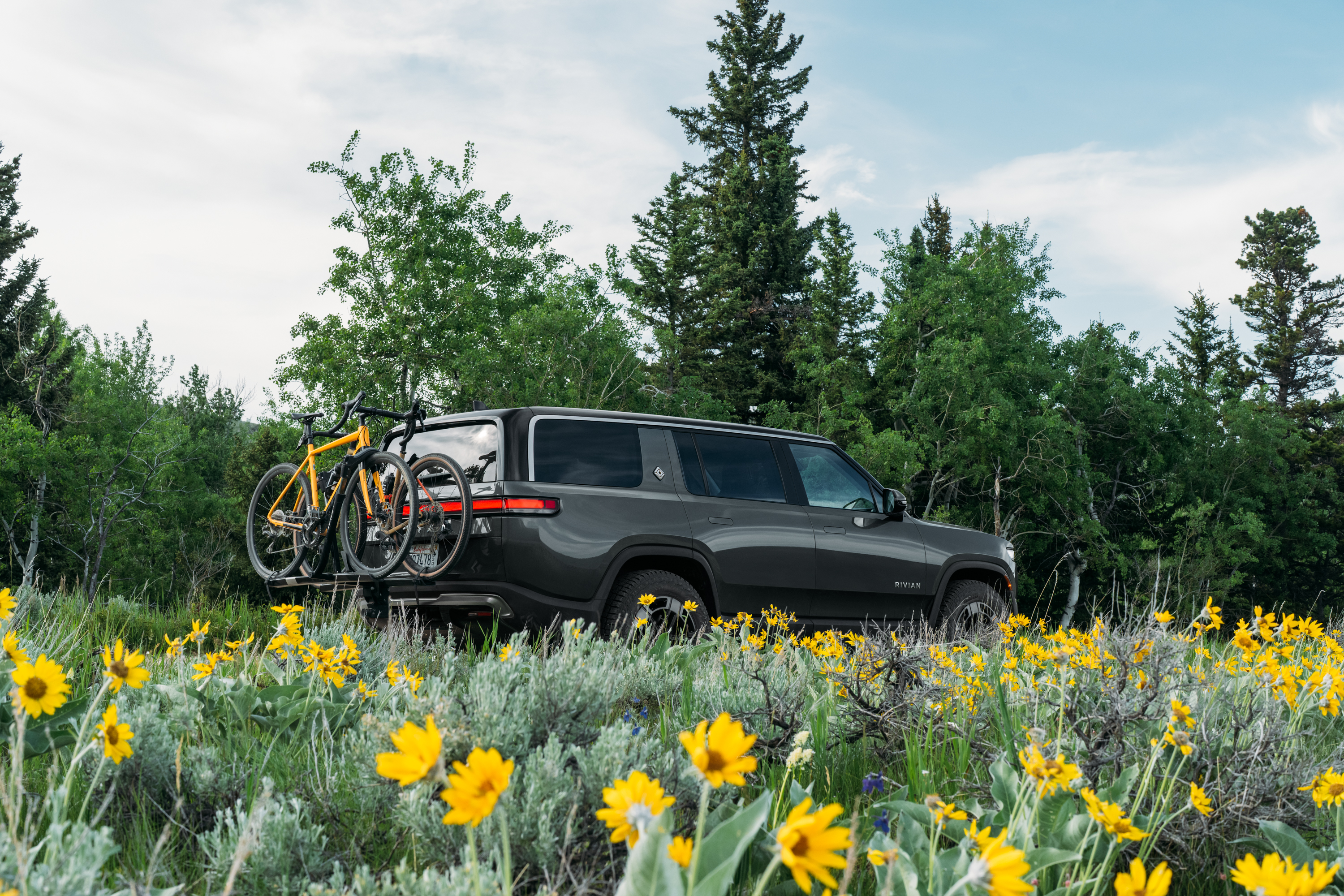 Rivian SUV parks in a meadow of yellow wildflowers. Two bicycles are ...