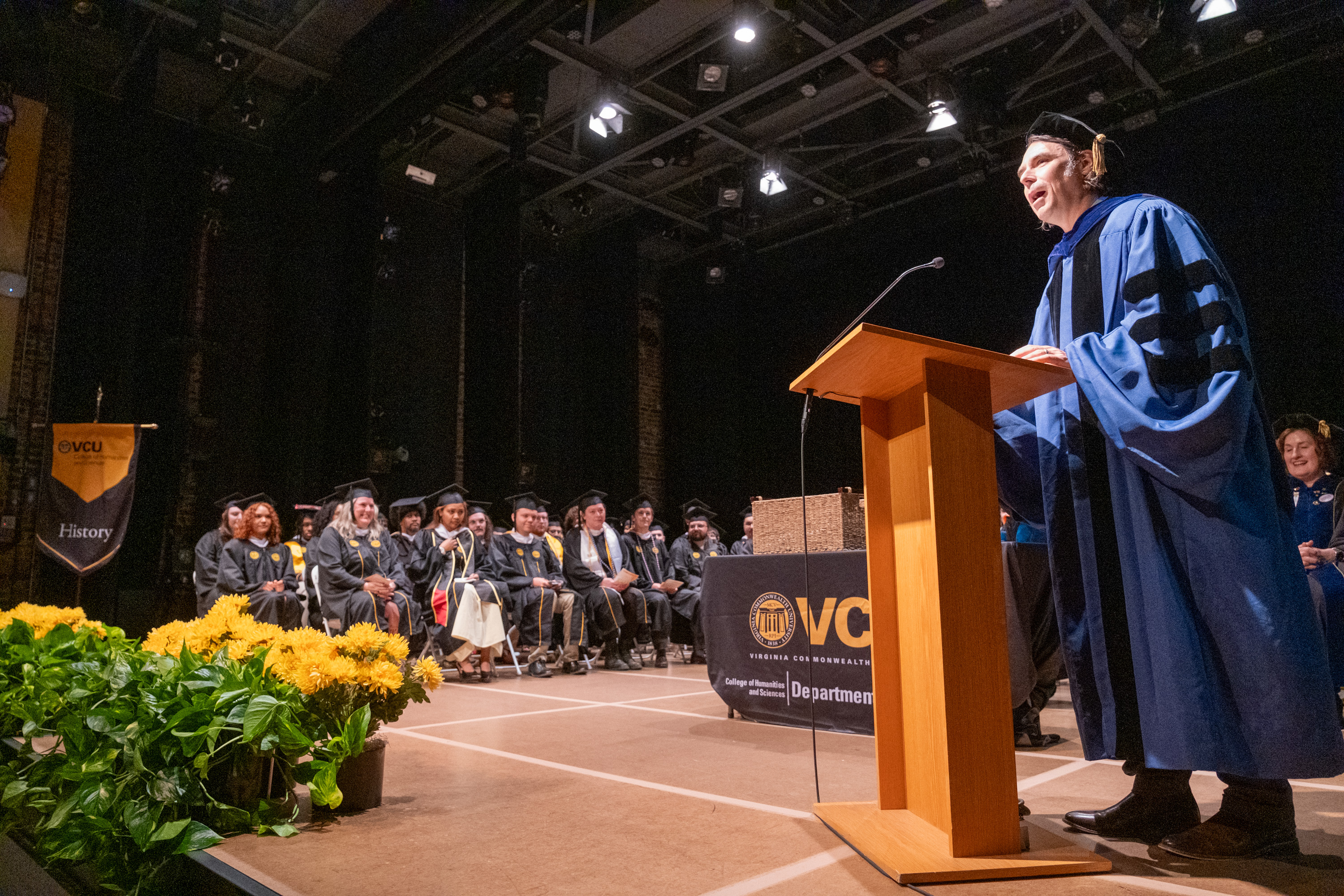 A speaker in blue gown stands at a podium addressing graduates seated ...