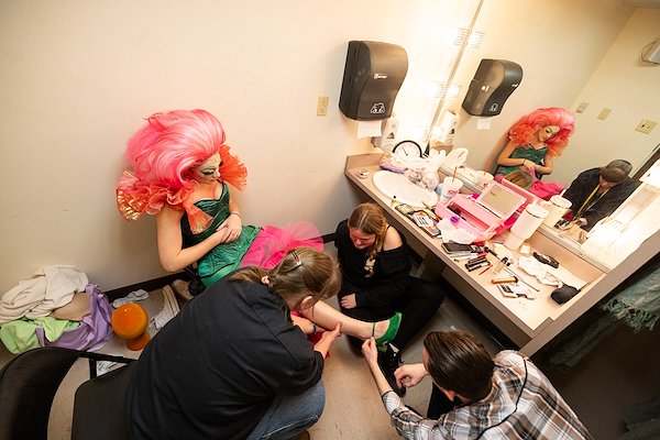 An actor in an elaborate pink puffy wig wearing a bright green costume sits in a chair in a dressing room in front of a mirror while three people crouch beside her adjusting her green shoe.