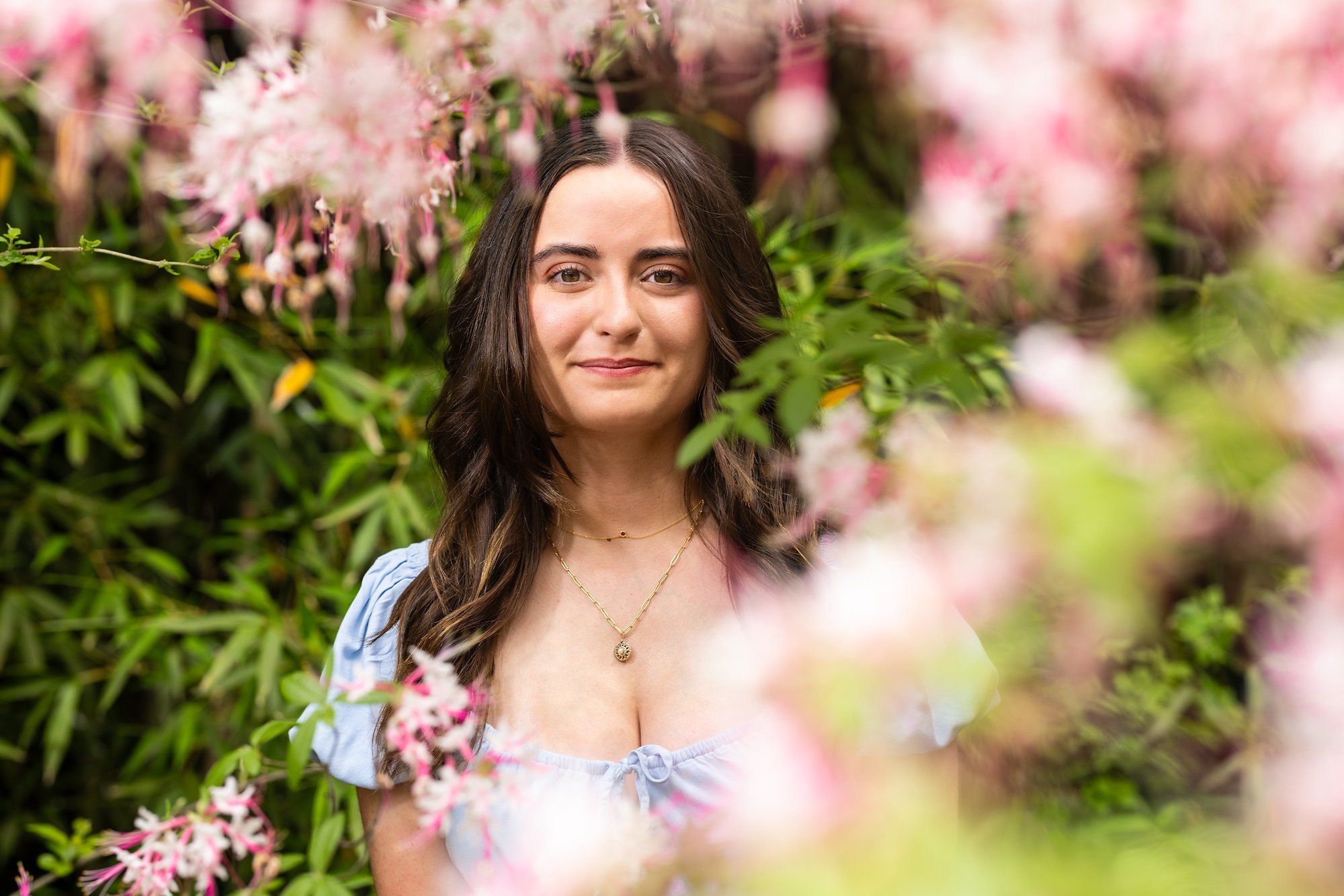 A white, woman with long dark, middle-parted hair and a small, closed lip smile gazes at the camera through a circle of pink flowers and greenery in a garden.