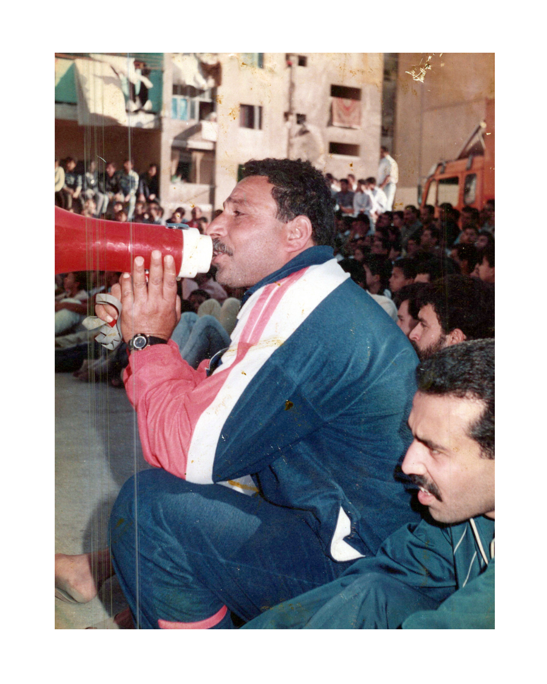 Man squats, speaking into red megaphone, wearing blue tracksuit with ...