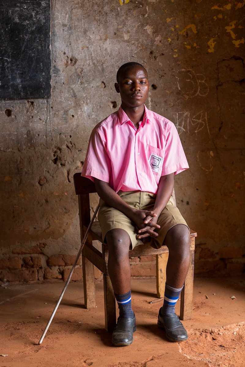 Portrait of Saleh, sitting on a wooden chair in his school uniform with his white cane resting against him.