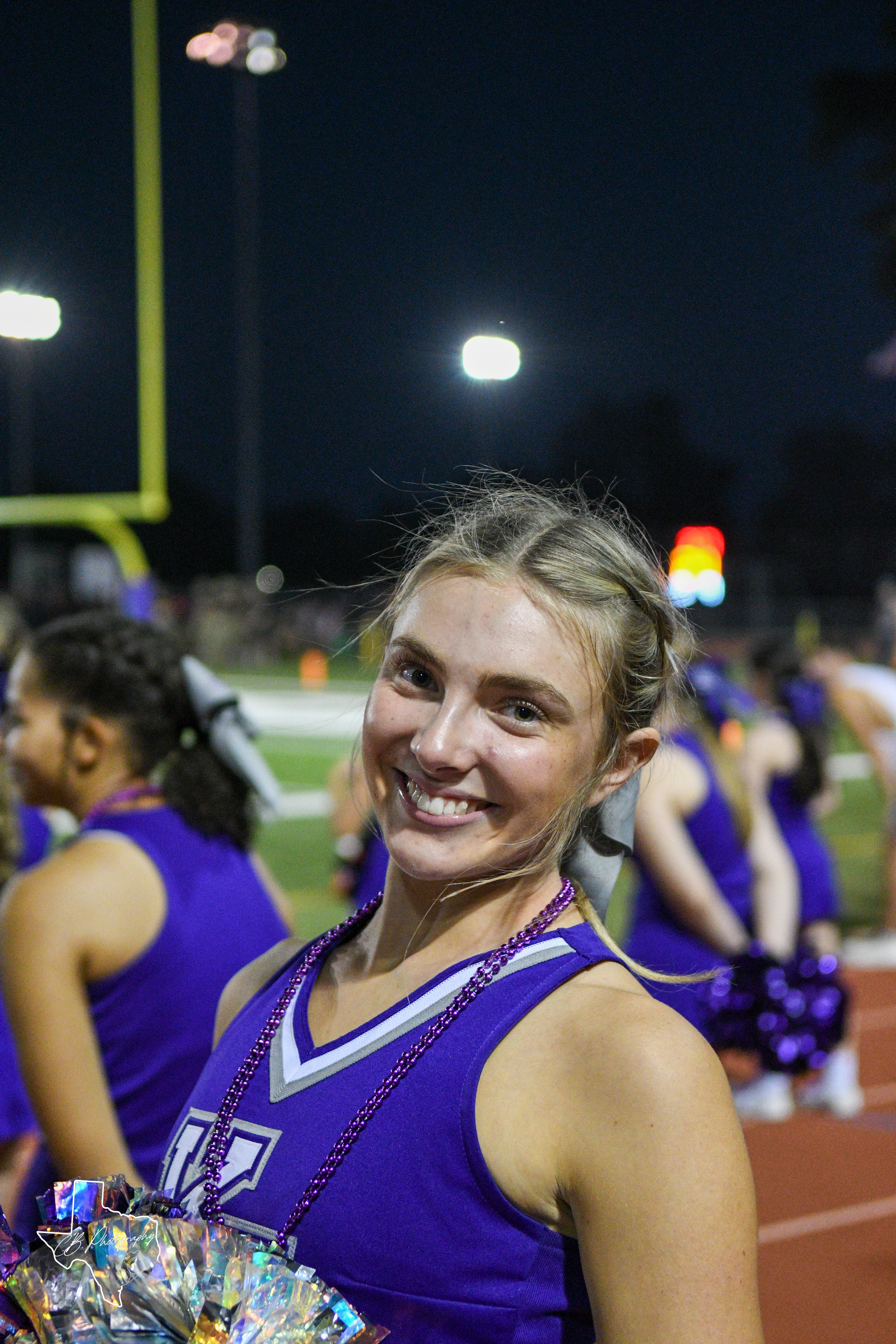 Smiling cheerleader in purple uniform holds pom-poms on a football ...
