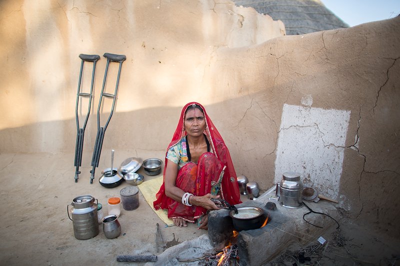 Manju crouching down, surrounded by cooking pots. Her crutches rest against a wall in the background.