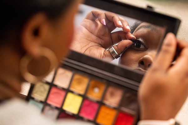 Closeup of a makeup palette with a young, Black women looking into the palette’s mirror as she applies a false eyelash.