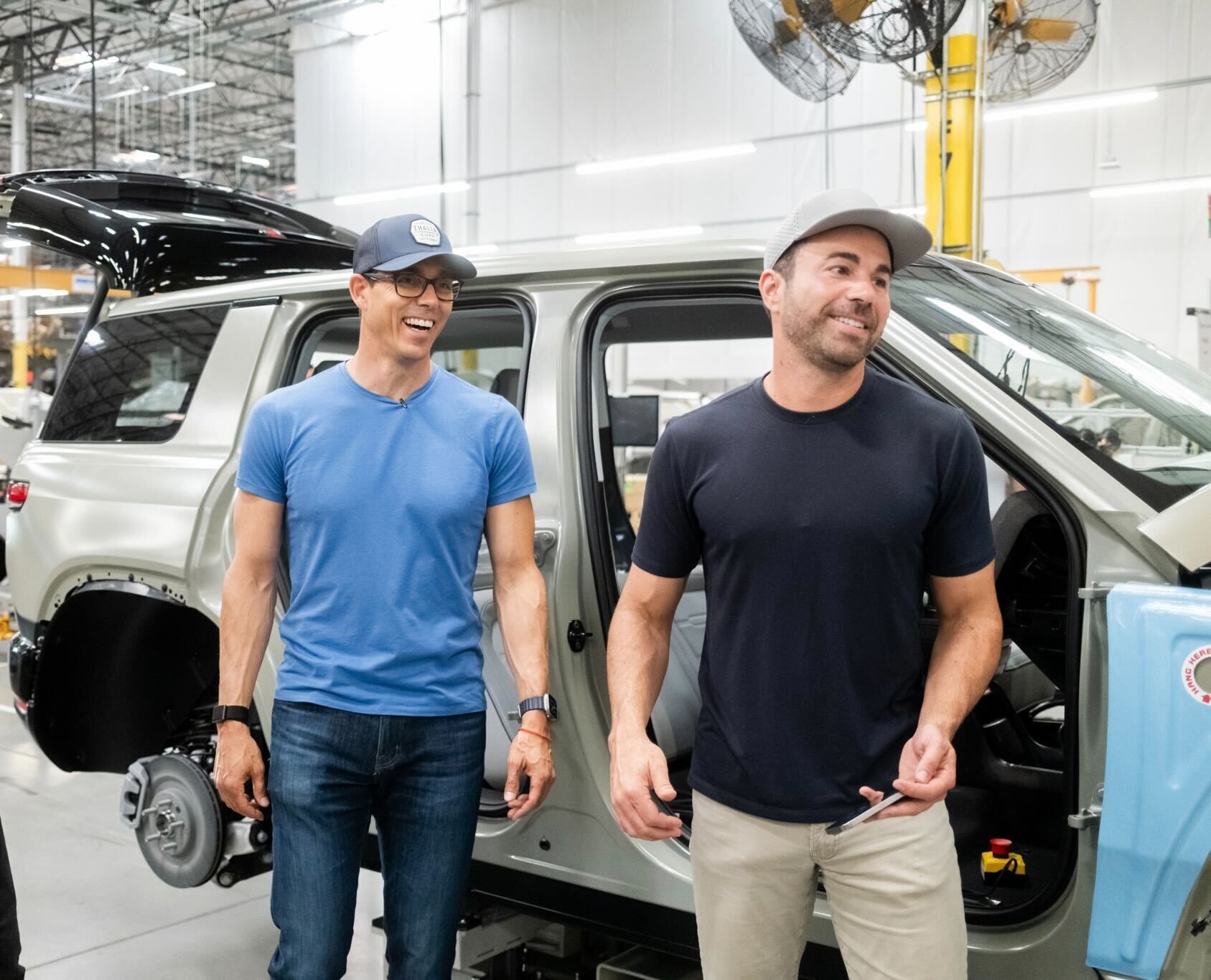 Two men stand smiling next to a Rivian vehicle in a factory setting ...