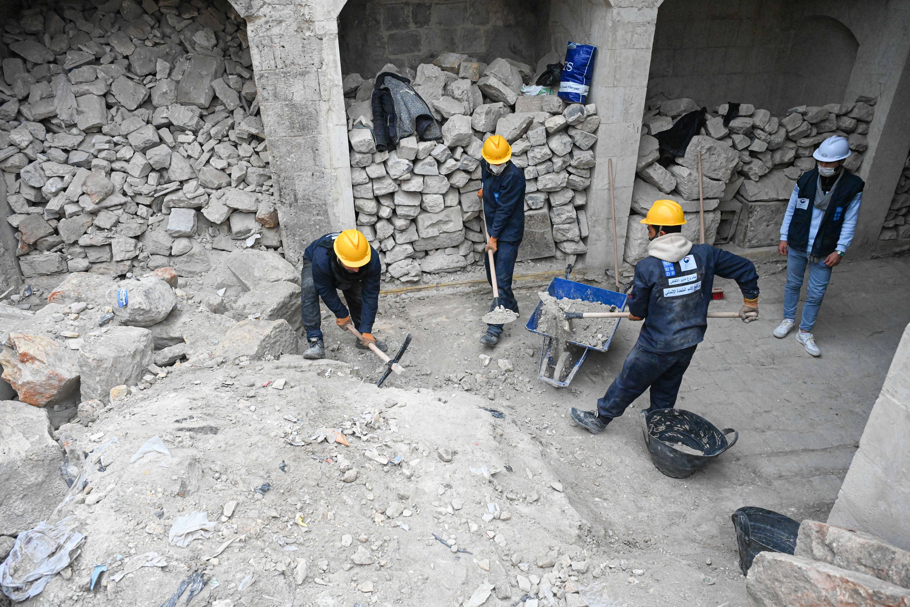 Workers in yellow hard hats clear rubble with shovels and a pickaxe, as ...