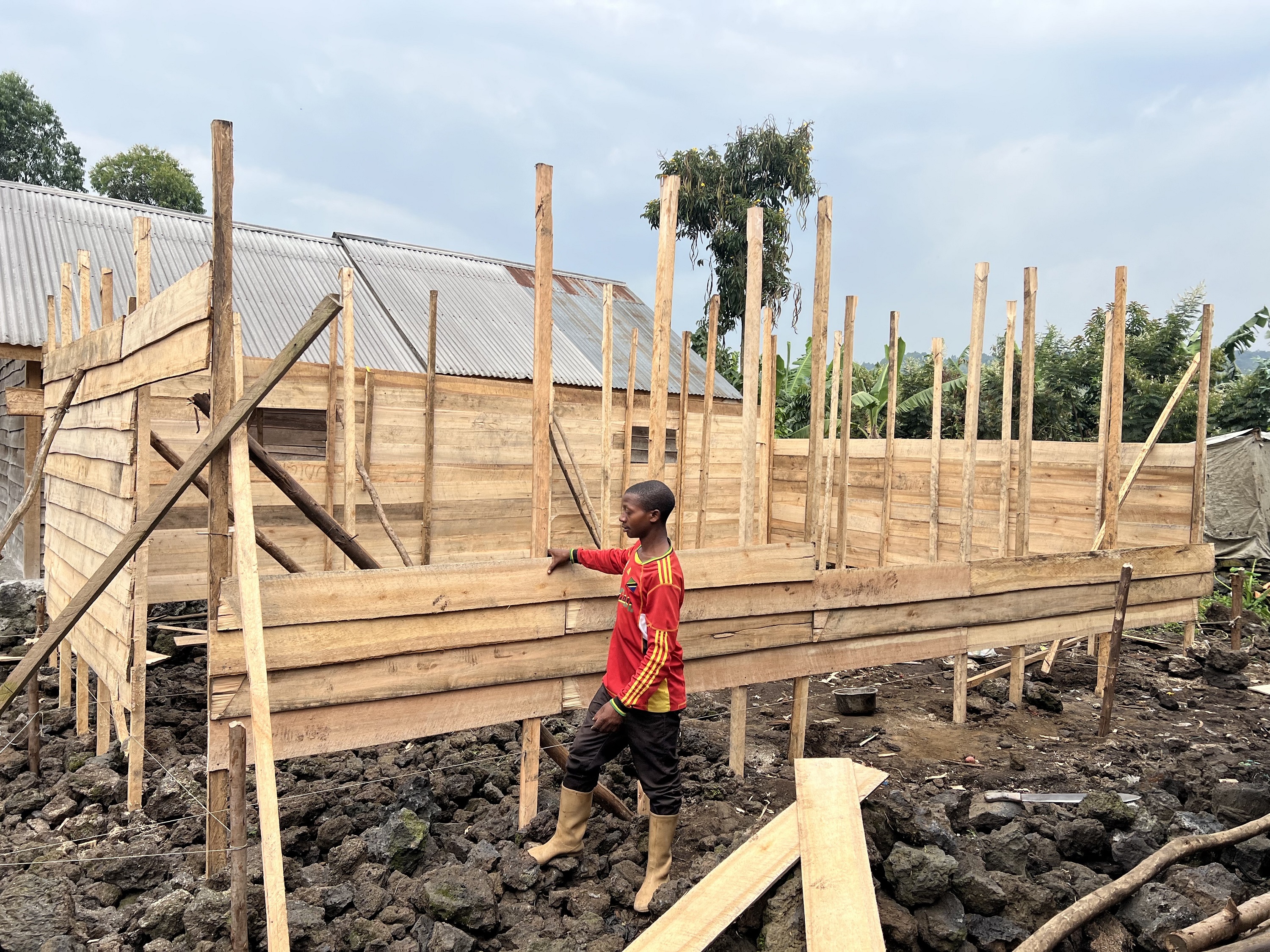 A boy stands near a wood-frame structure under construction. He wears a ...