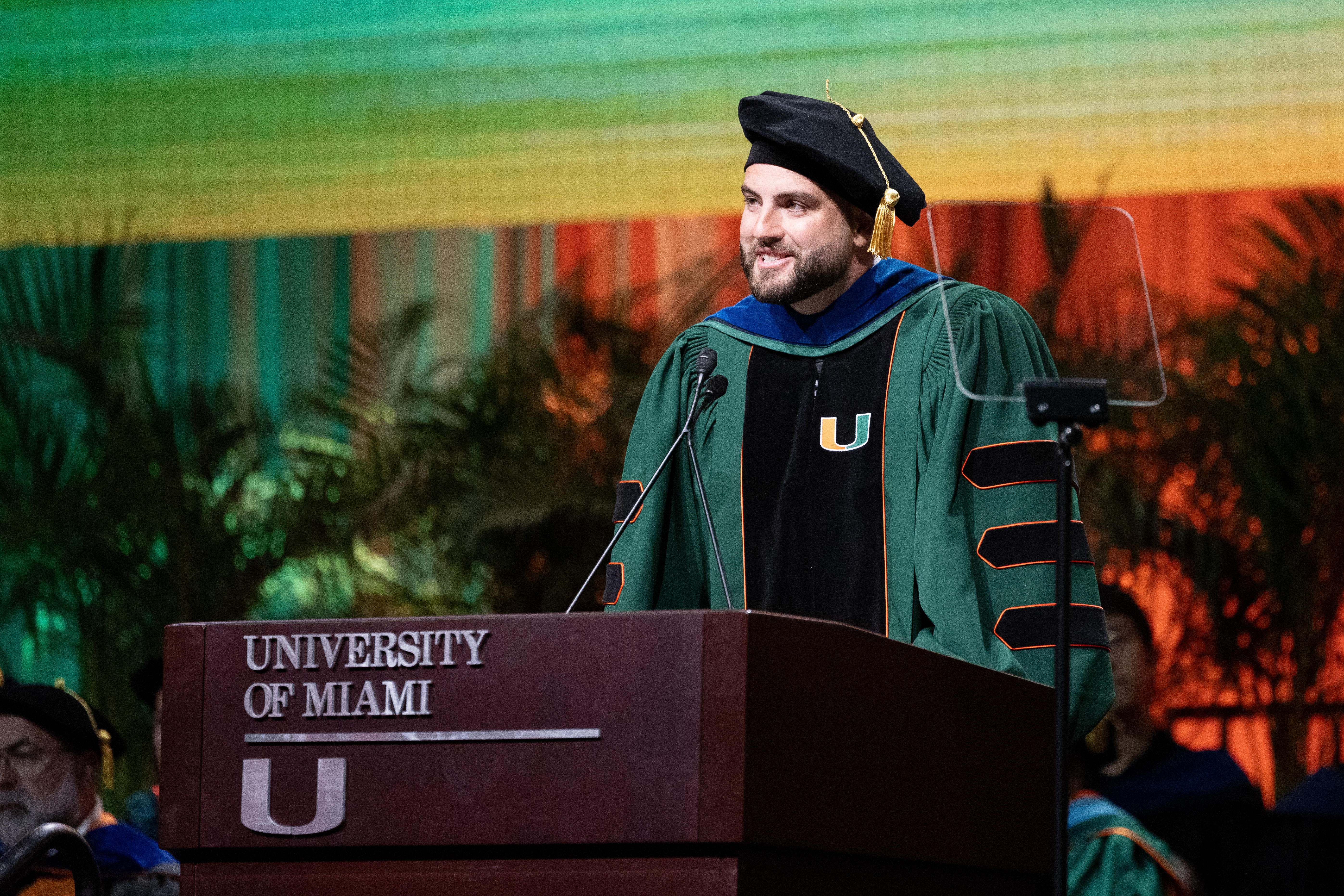 Man in graduation gown speaks at podium for