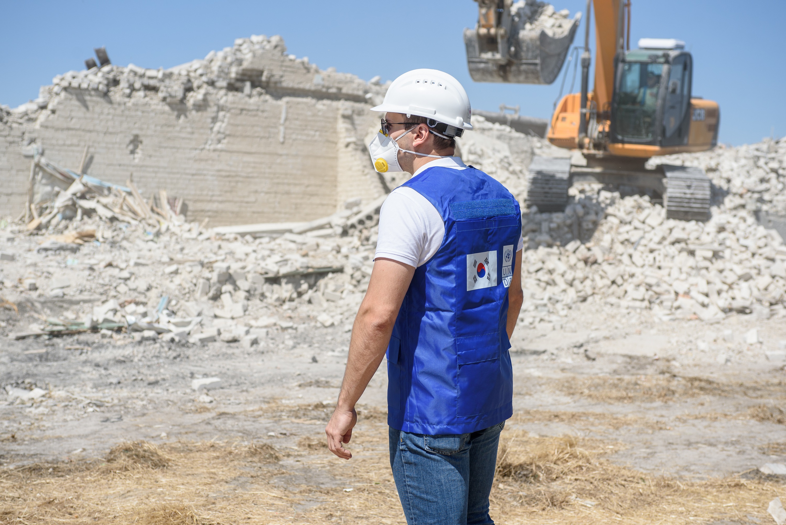A man wearing a hard hat and mask stands in a debris field, looking ...