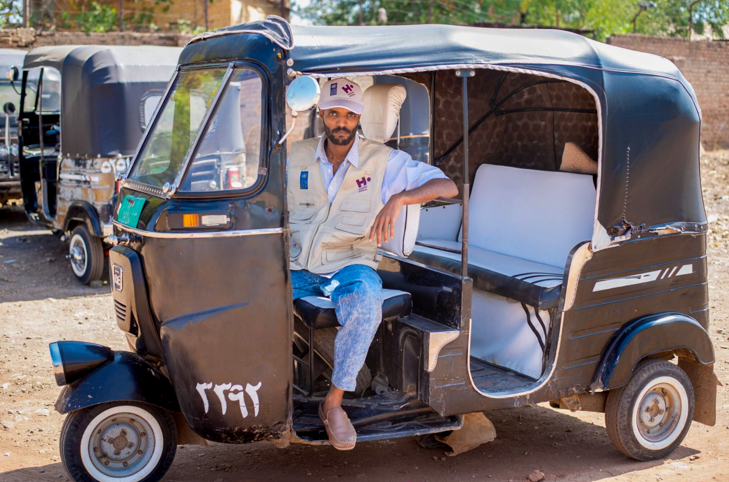 Man in a vest and cap sits in a black auto rickshaw, arm resting on the ...