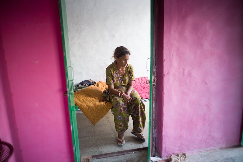 Photograph taken through a doorway of Dallu sitting on her bed, with her hands resting in her lap.