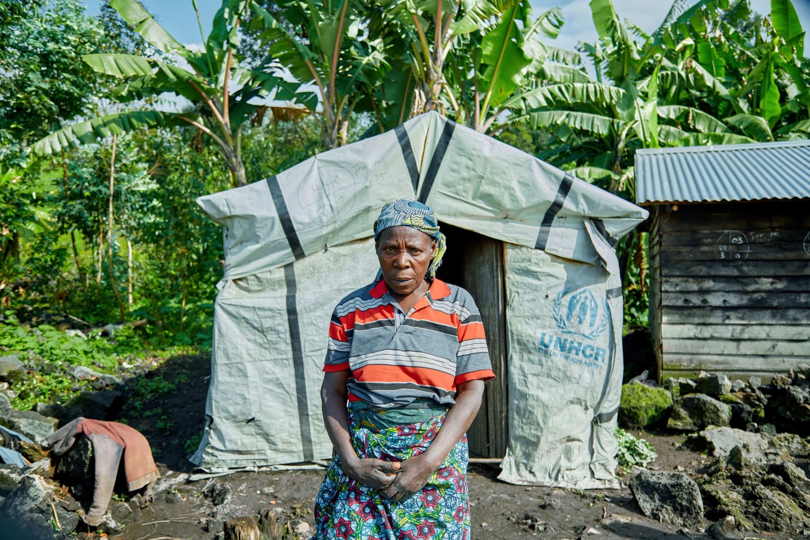 An older woman stands in front of a UNHCR tent in a rural area. She ...