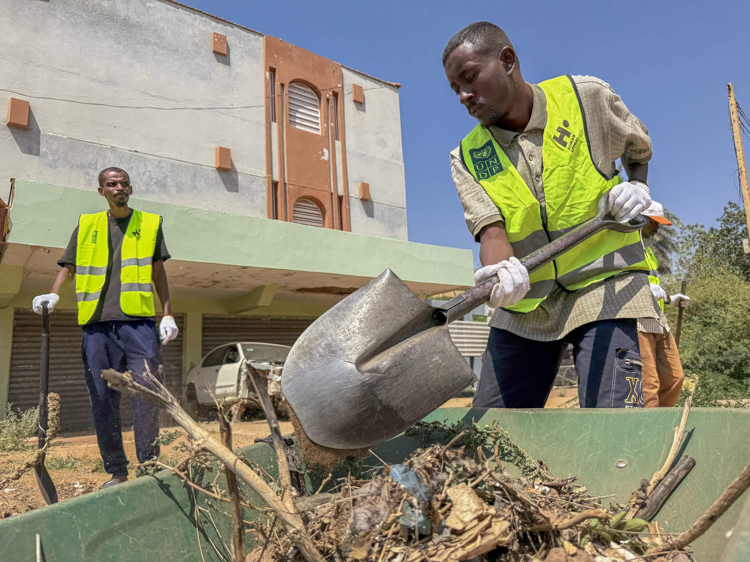 Two men wearing neon vests clean debris. One man shovels debris into a ...
