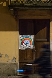 In the quiet of the night, a cyclist passes a handmade rainbow banner bearing an Italian phrase meaning "It will all be well." The banners are appearing around Cremona during the COVID-19 crisis.