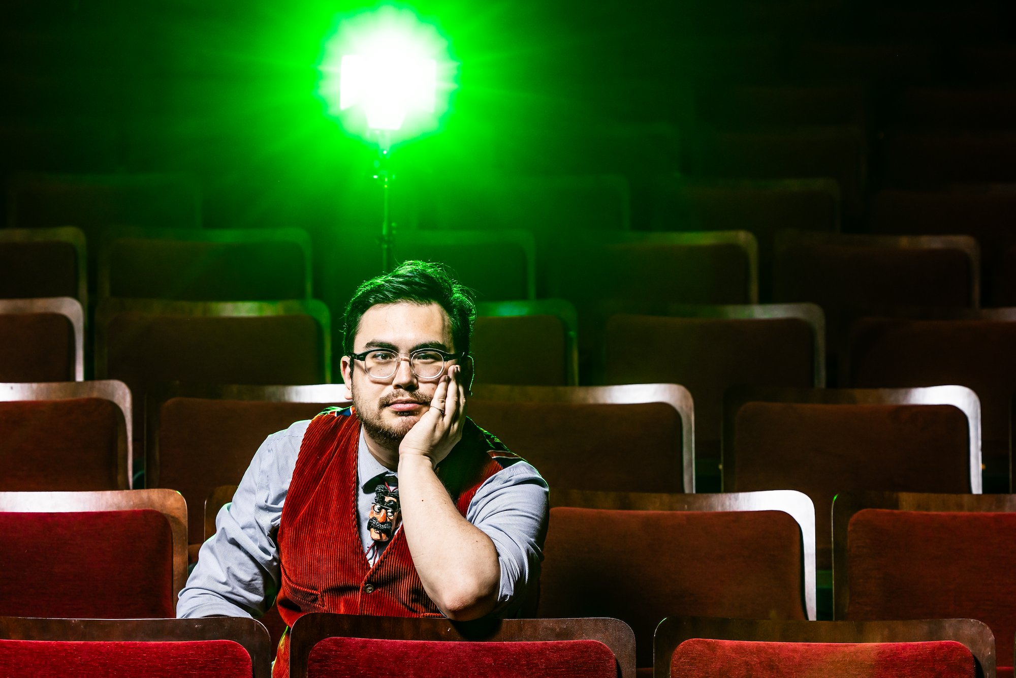 A Latino man with short, dark hair sits in an auditorium wearing horned rim glasses, and a red vest over a long-sleeved blue shirt. His chin is propped up on his hand and a green spotlight shines behind him.