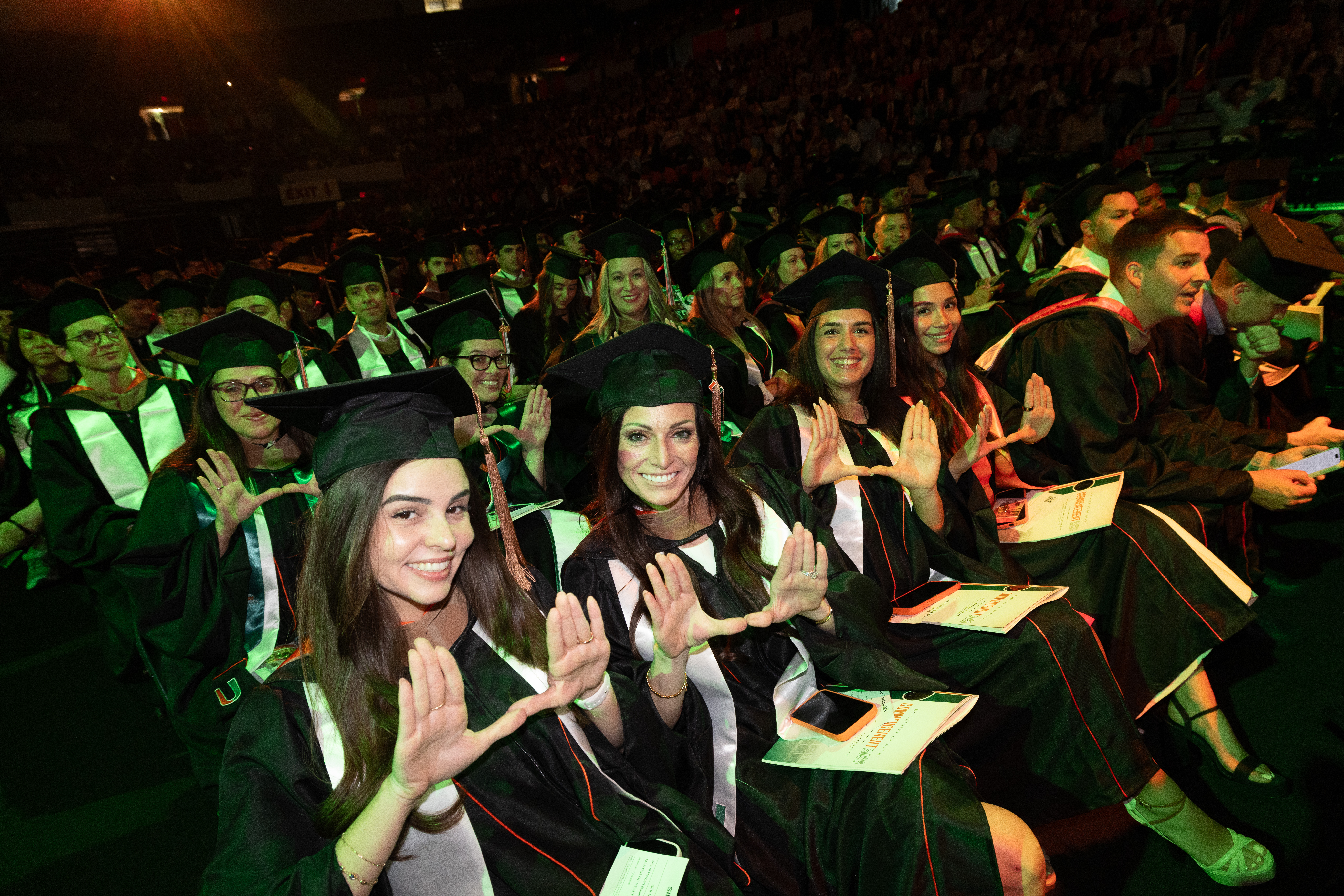 Graduates in black gowns and caps sit in rows, smiling and holding up ...
