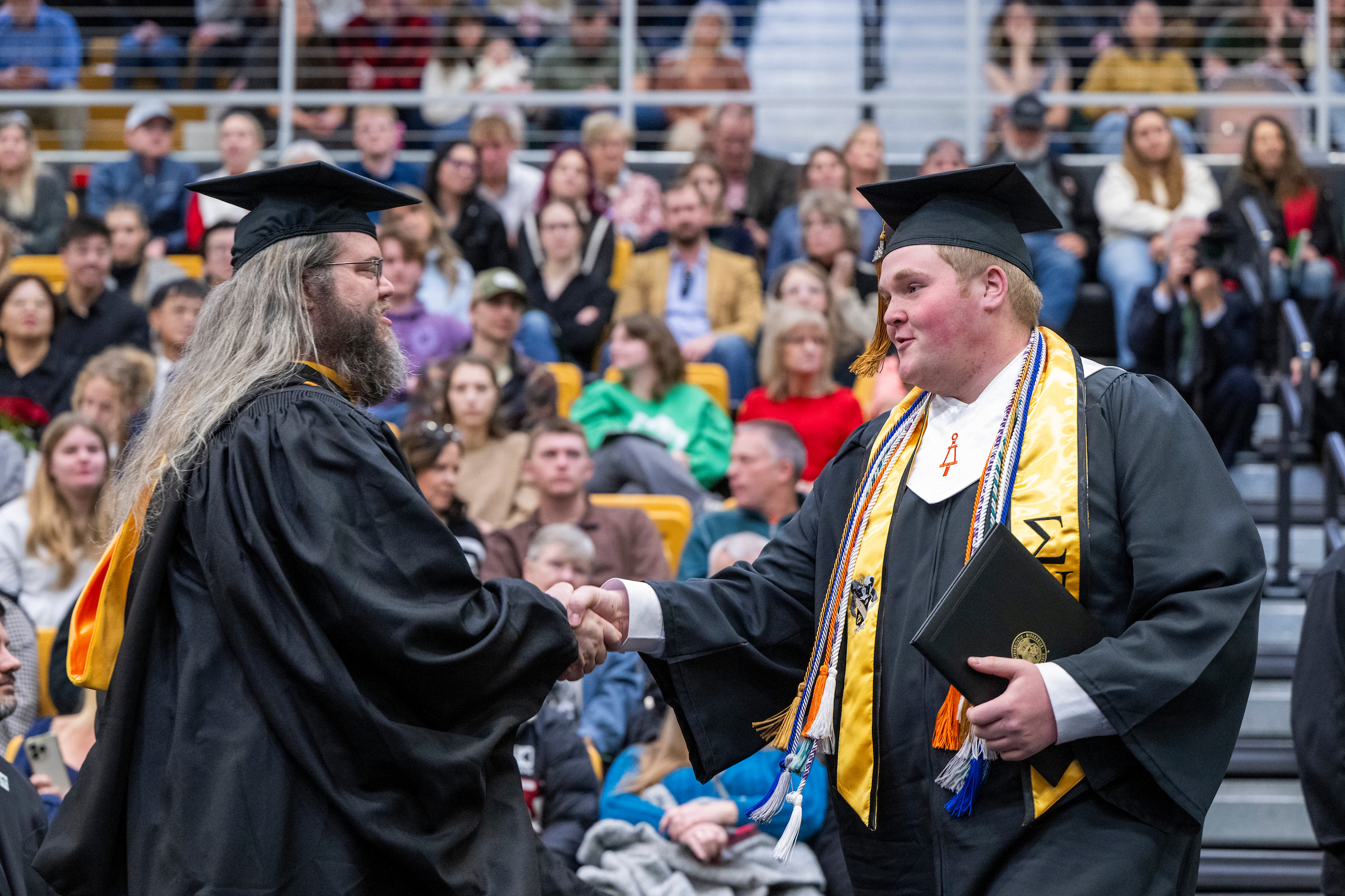 Two graduates in black gowns shake hands in front of a crowd. One ...