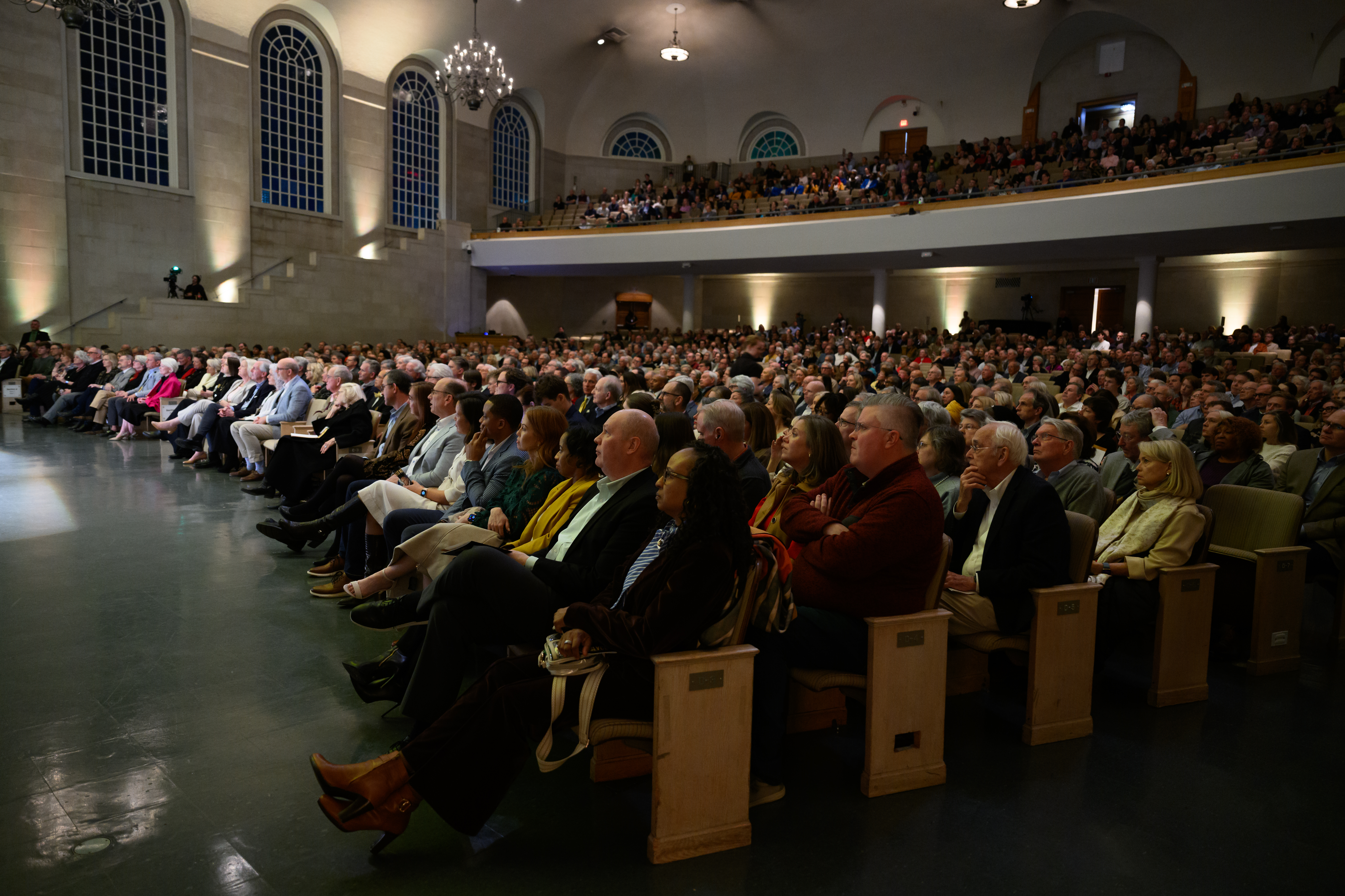 Audience members sit in rows of wooden chairs in a large auditorium ...