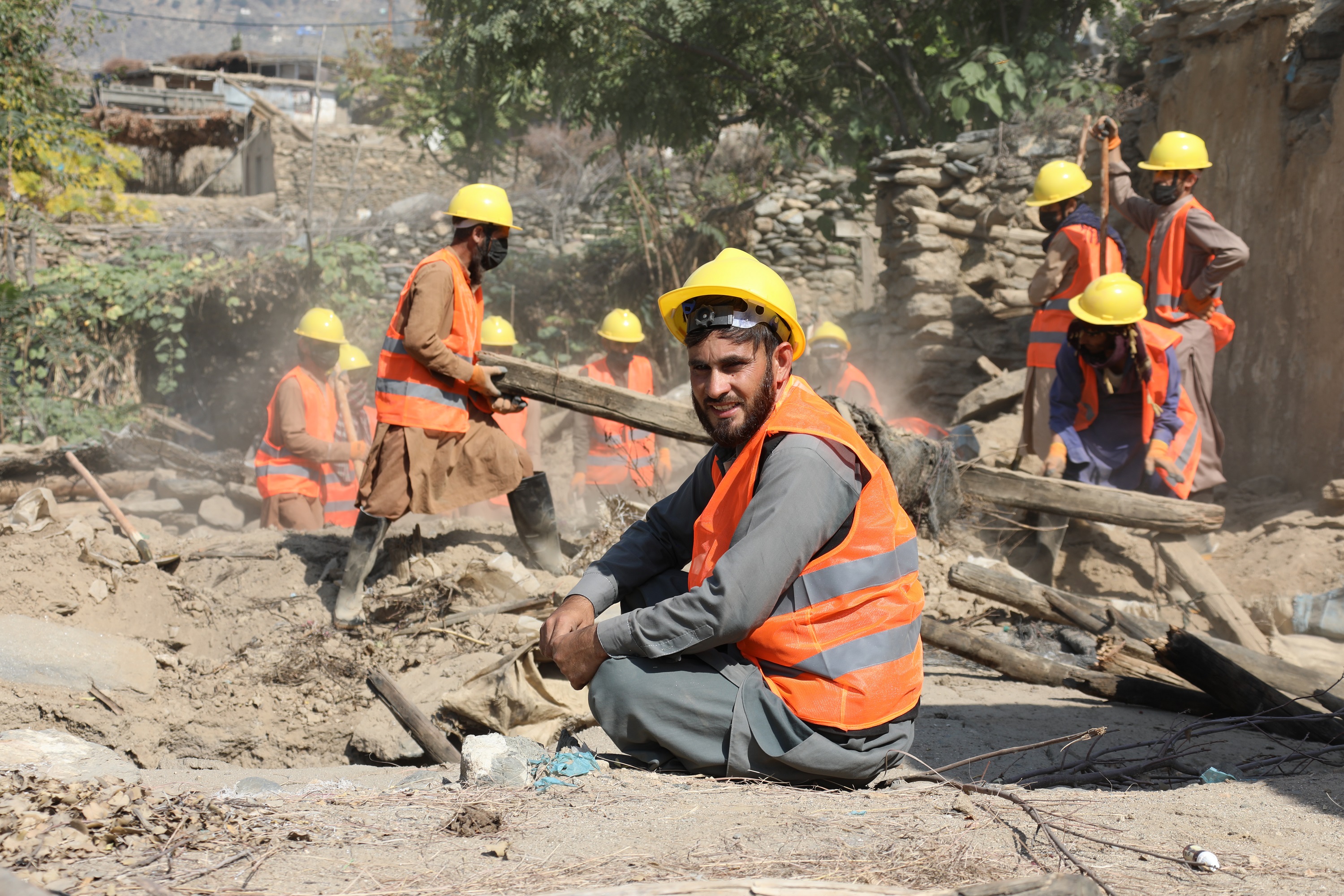 Man in hardhat and safety vest smiles, sitting amid rubble, with rescue ...