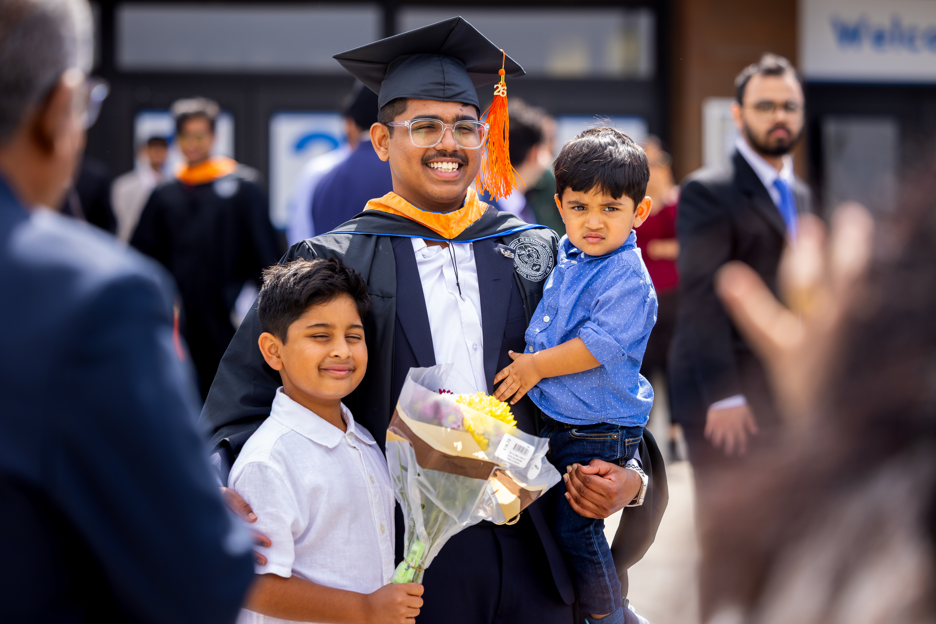 A graduate smiles, wearing cap and gown, holding a toddler and standing ...