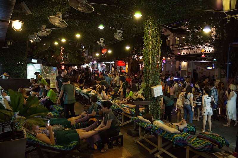 Tourists enjoy cheap foot massages alongside a busy street of bars, restaurants, and street food vendors.
