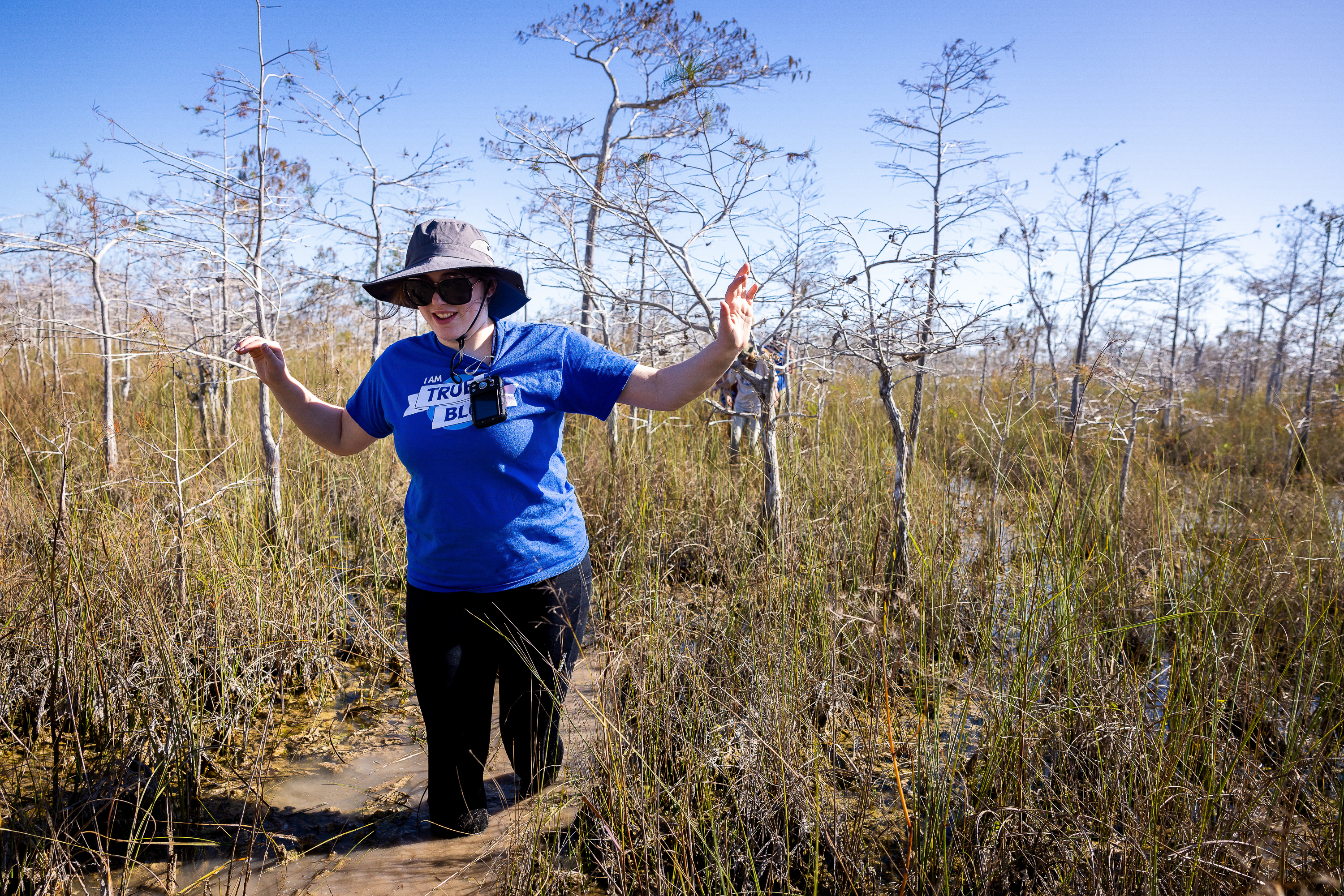 A person wearing a hat walks through a swamp area with water up to ...