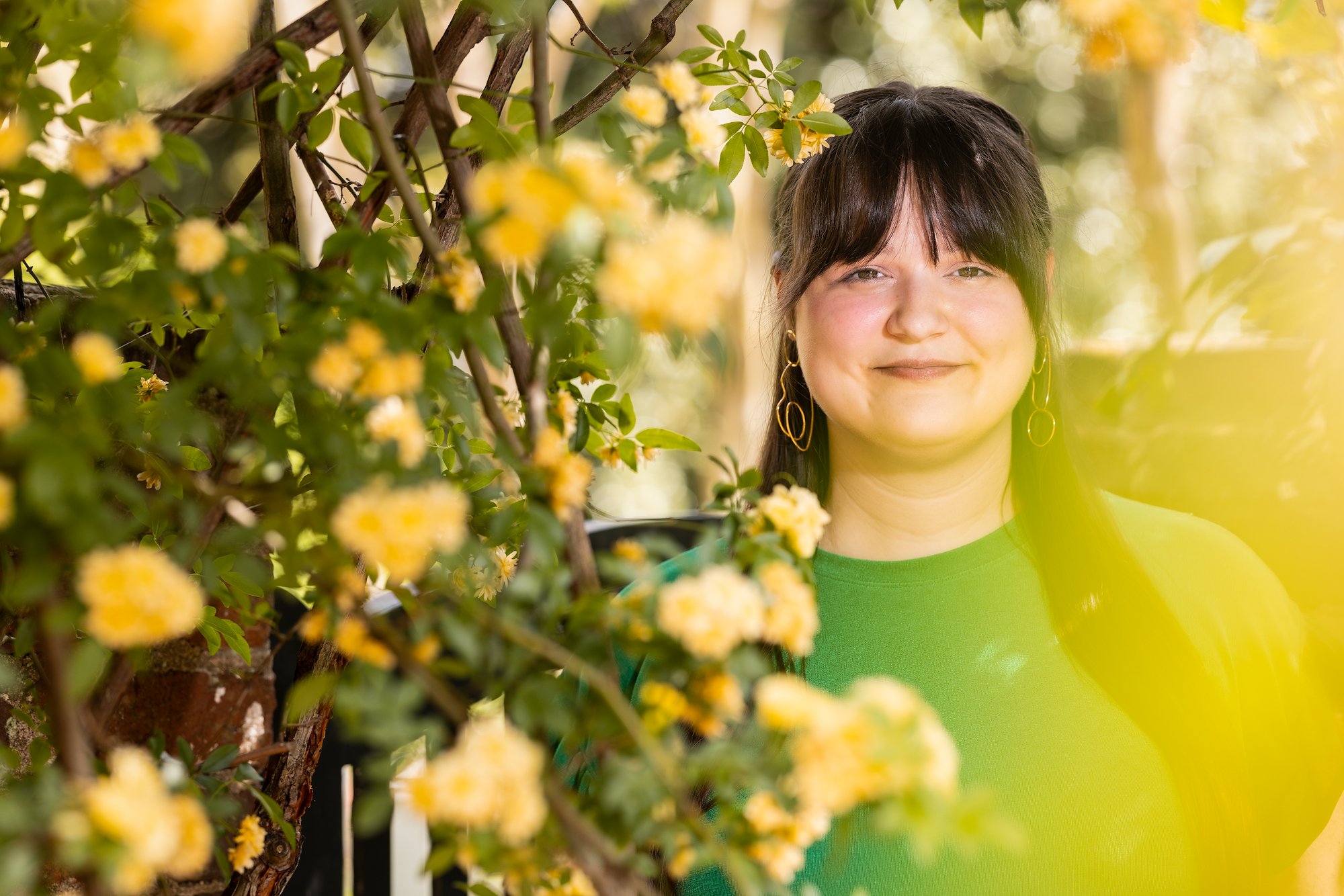 A white woman with dark bangs and long dark hair is wearing a green shirt and dangly gold earrings as she stands peeking out of a host of yellow flowers in a garden.