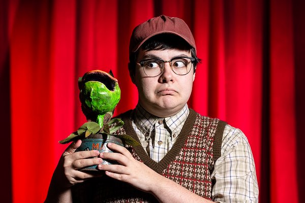 A white person wearing a tweed ball cap, horned rim glasses, a button-down shirt and a brown and orange V-neck vest holds up a fake potted plant that looks like a Fly Catcher, which was created for the play “Little Shop of Horrors.” His expression is one of worry and he stands in front of a red curtain.
