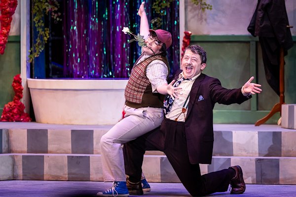 On a stage an actor playing Seymour holds a rose in his mouth and sits on the bended knee of an actor playing Mr. Mushnik. Both have their arms flung out with jazz hands.