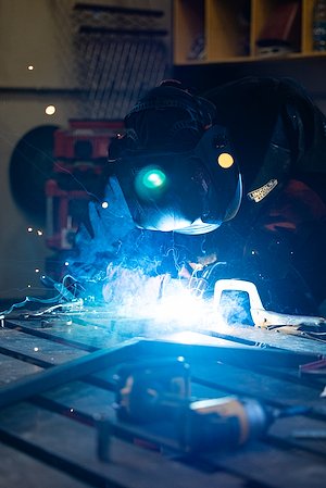 A man in a welding mask leans over a piece of metal. Blue sparks fly around him.