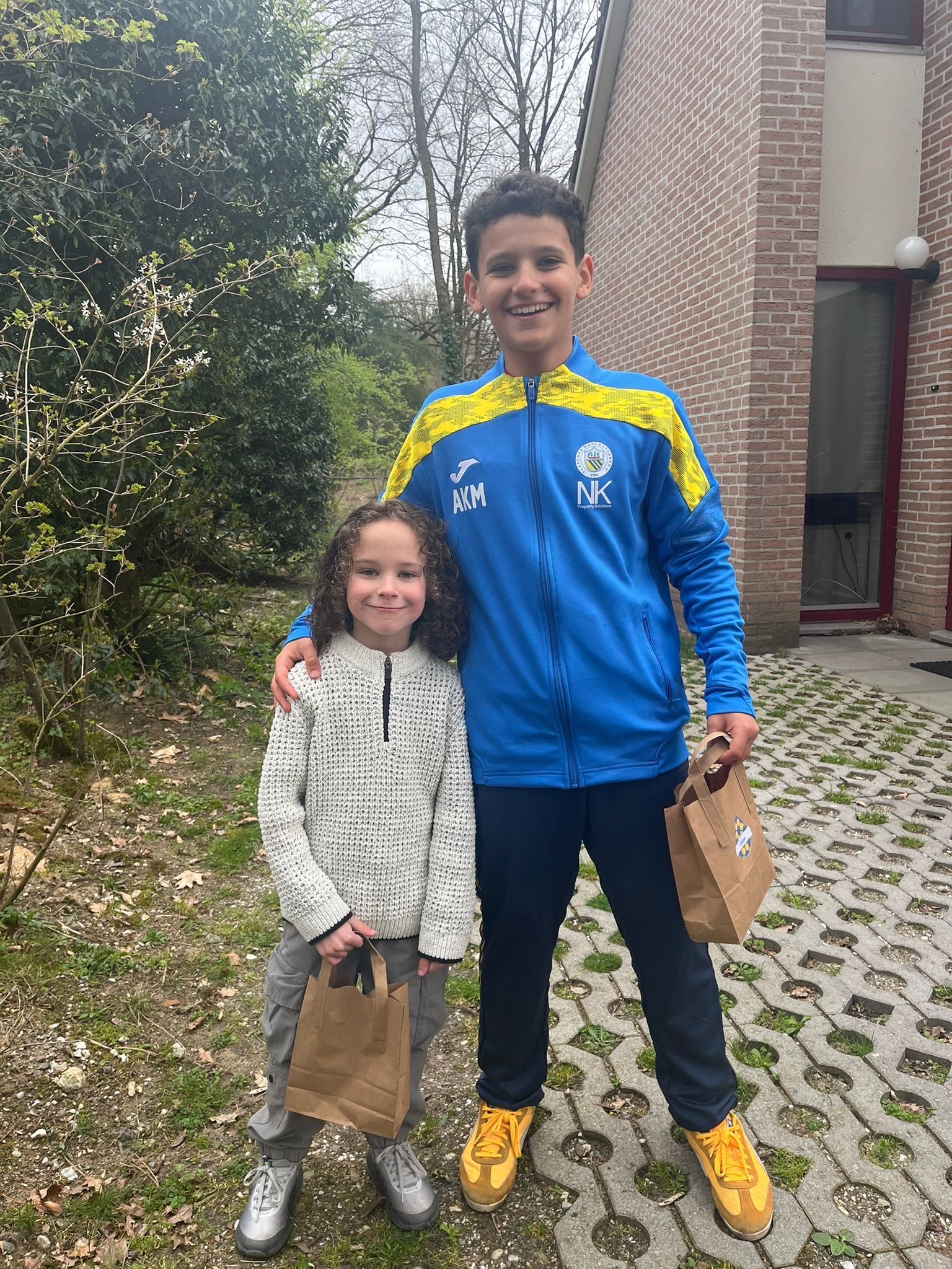 Two smiling boys stand outside, holding brown paper bags. The older boy ...