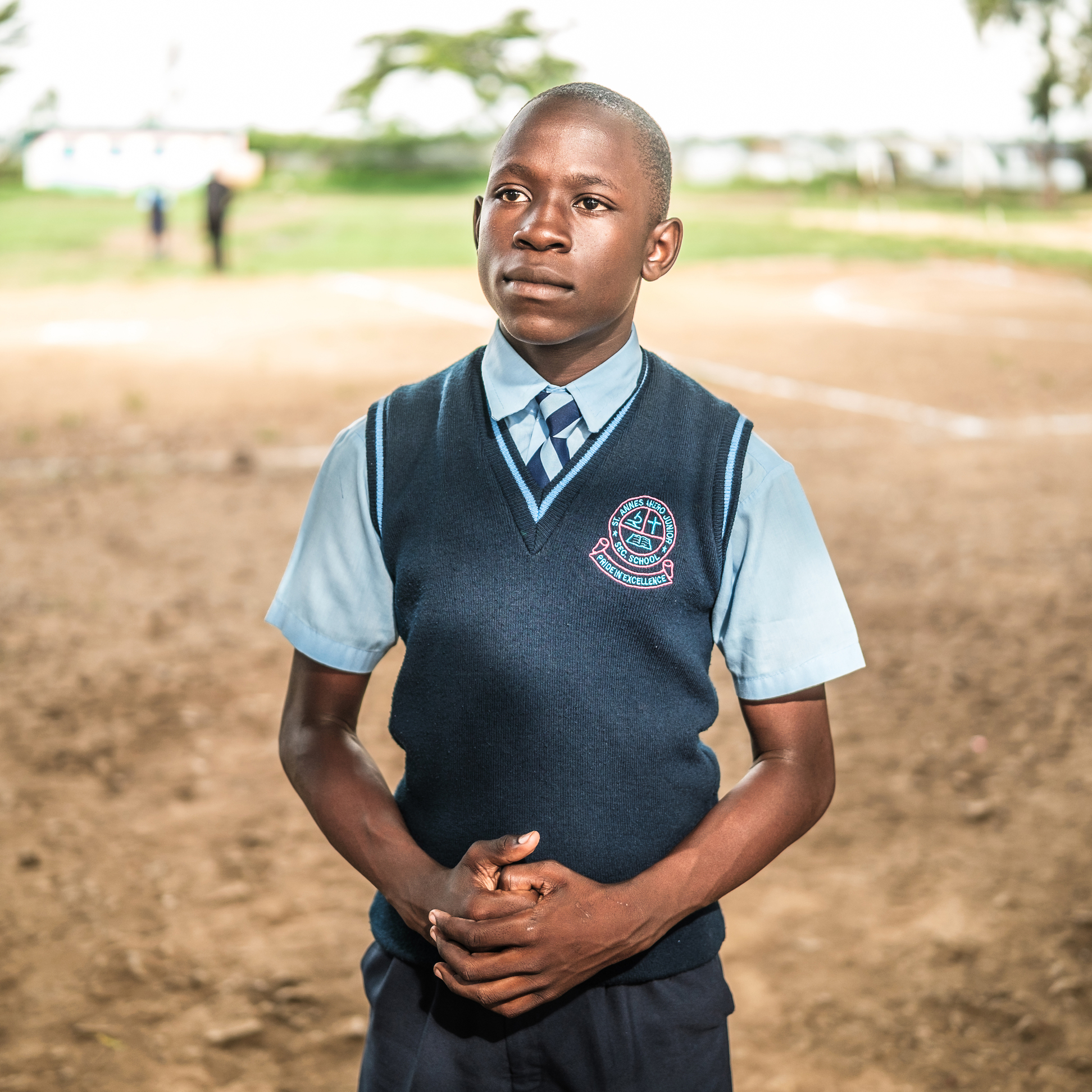 Kenyan schoolboy in uniform stands on a dirt field. He wears a blue ...