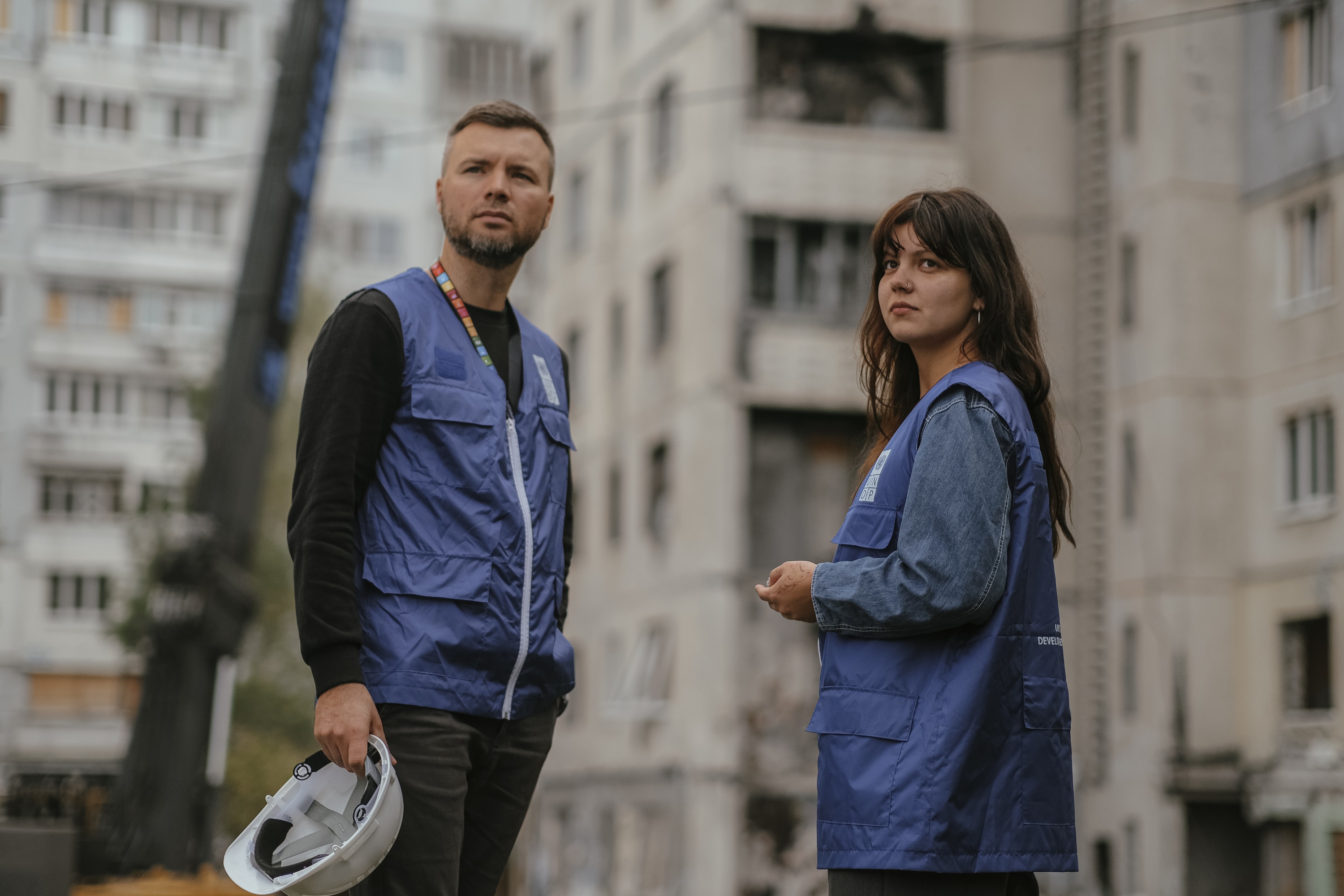 Two aid workers stand in front of a damaged building. The man holds a ...