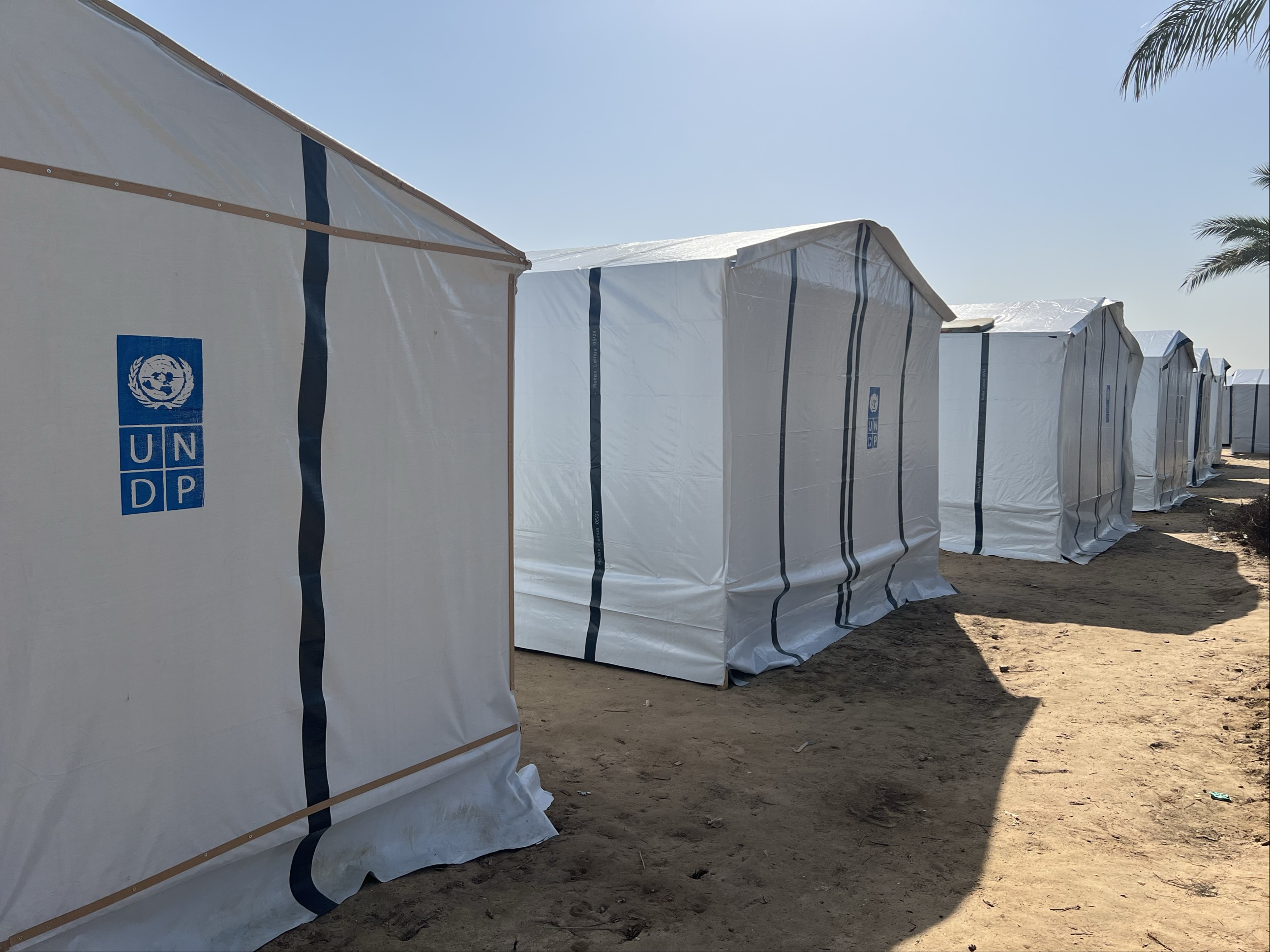 Row of white temporary shelters with the UNDP logo, on a sandy lot ...