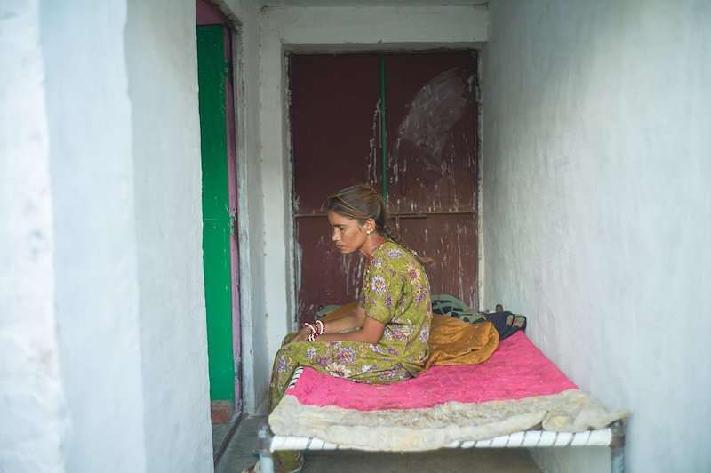 Profile photograph of Dallu sitting on her bed in a sparsely furnished room.