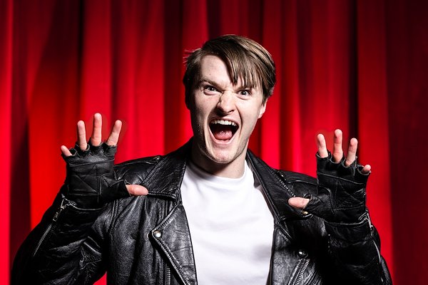 A white, blonde man wearing a leather jacket, black leather, fingerless gloves and white T-shirt holds up his hands like fangs and snarls at the camera. A red curtain is the backdrop.