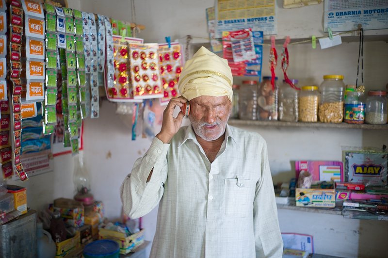 Sankarlal standing in his shop, speaking on a mobile phone. He is surrounded by brightly coloured objects for sale.