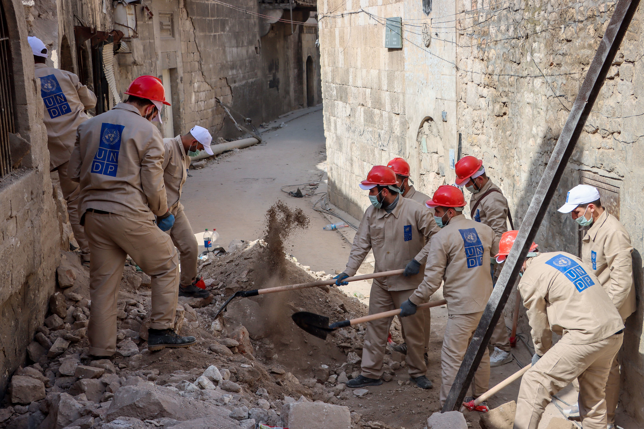 UNDP workers wearing hard hats and face masks shovel rubble in a narrow ...